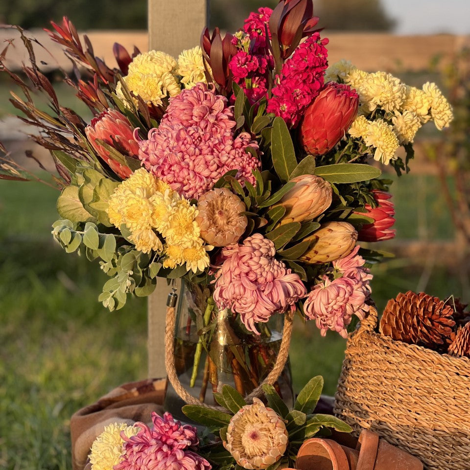 A glass vase containing a colorful bouquet of flowers including chrysanthemums and other seasonal flowers, arranged outdoors with a basket and wooden elements in the setting.